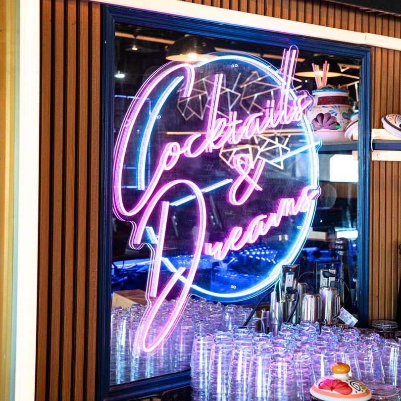 Neon sign in a window at Chako's Social reads "Cocktails & Dreams" above rows of glasses.