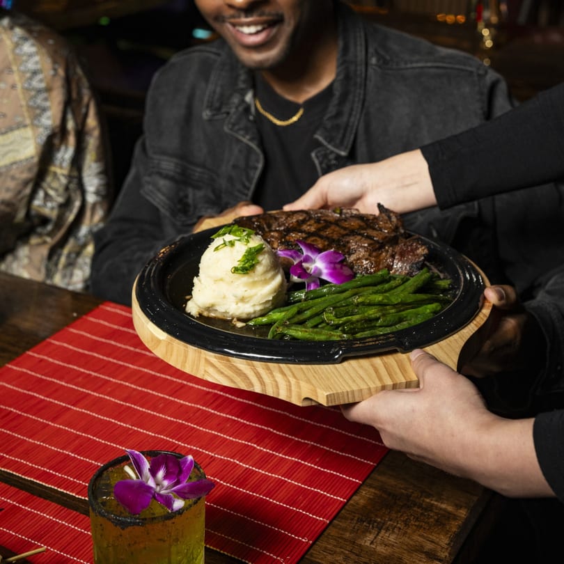 A server delivers a sizzling steak with mashed potatoes and green beans to a smiling customer.