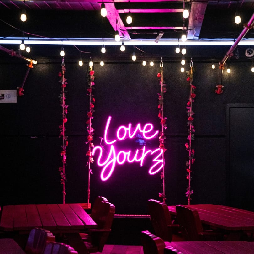 A neon pink "Love Yourz" sign hangs between two rose-covered swings, above red picnic tables.